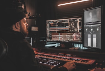 A person wearing a beanie is seated at a desk, working on digital audio workstations displayed on multiple computer monitors. The room appears to be a studio with soundproof walls, and the environment is dimly lit with a warm ambiance from overhead lighting. The screens show complex audio editing software with various tracks and controls.