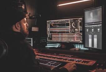 A person wearing a beanie is seated at a desk, working on digital audio workstations displayed on multiple computer monitors. The room appears to be a studio with soundproof walls, and the environment is dimly lit with a warm ambiance from overhead lighting. The screens show complex audio editing software with various tracks and controls.