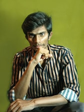 A person with a thoughtful expression is pictured wearing a striped shirt. The background is a solid, muted yellow-green color. The lighting highlights their facial features, suggesting deep thought or introspection.