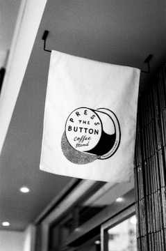 A monochrome image featuring a fabric sign hanging from a bracket. The sign displays a round logo with the words 'Press the Button Coffee Stand', surrounded by artistic shading.