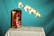 A still life setup featuring a metal fuel canister labeled 'TRUFUEL' with a 50:1 mix ratio, placed on a white cloth-covered table. A delicate branch of white orchids with purple centers stretches outwards from the canister against a solid turquoise background.