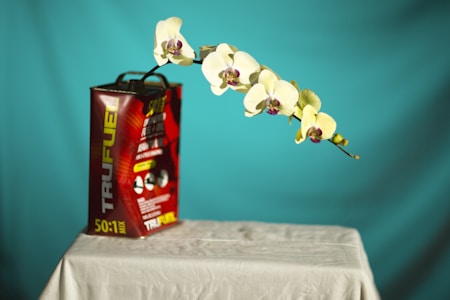A still life setup featuring a metal fuel canister labeled 'TRUFUEL' with a 50:1 mix ratio, placed on a white cloth-covered table. A delicate branch of white orchids with purple centers stretches outwards from the canister against a solid turquoise background.