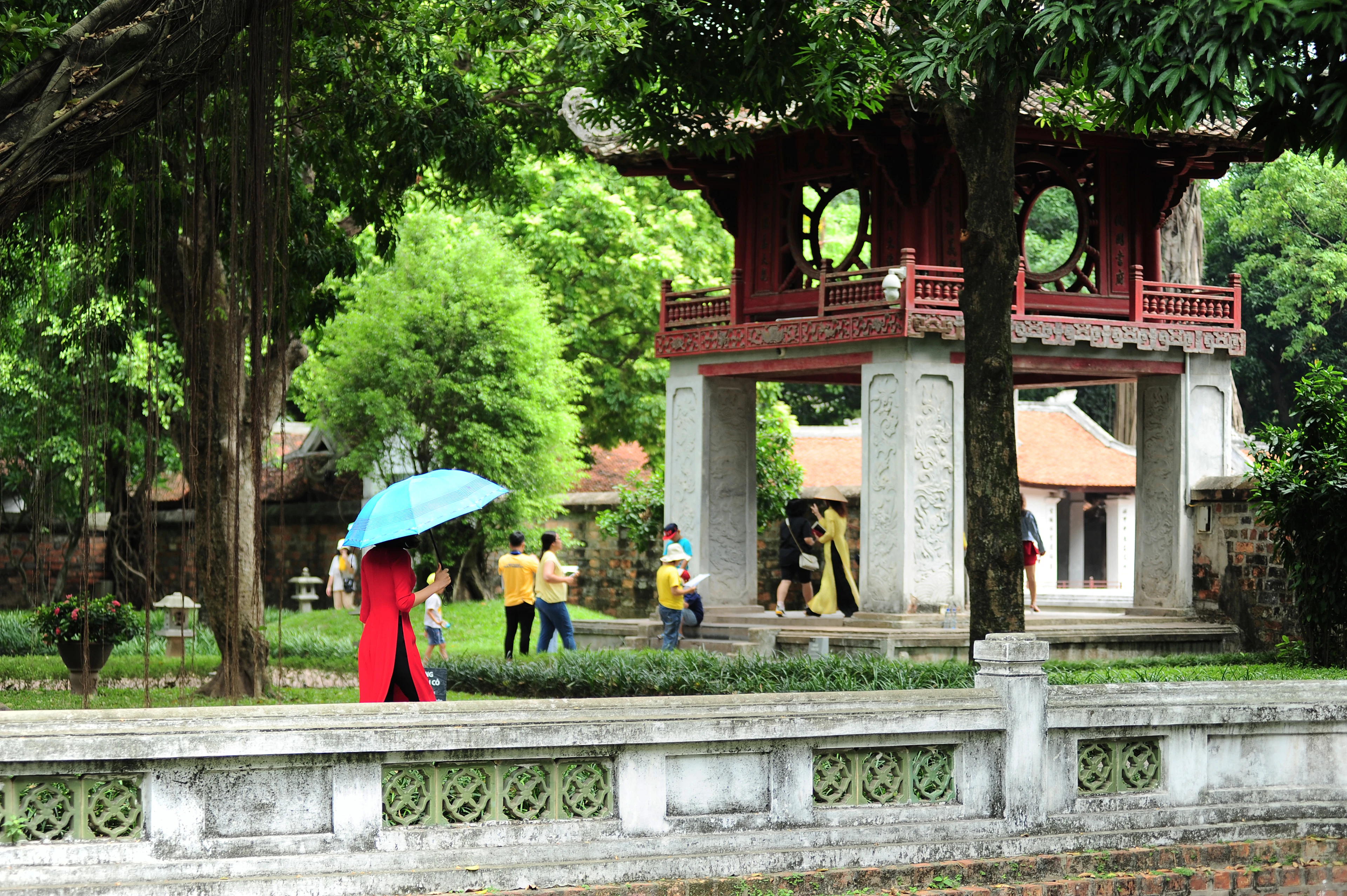 A woman in a red ao dai with a blue umbrella walks past the red Khue Van Cac pavilion.