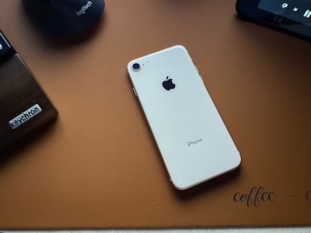 A light-colored iPhone is placed on a brown textured desk mat. To the left, there's a wooden keyboard or keyboard accessory labeled 'Keychron' and above it, a dark-colored Logitech computer mouse. Part of a tablet or another electronic device is visible in the top right corner.