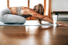 A person in athletic wear is performing a yoga pose on a mat indoors, with long hair falling forward. A tube of product labeled 'fringe mysa' is positioned nearby. The environment is well-lit with wooden flooring and a window in the background.