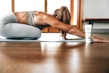 A person in athletic wear is performing a yoga pose on a mat indoors, with long hair falling forward. A tube of product labeled 'fringe mysa' is positioned nearby. The environment is well-lit with wooden flooring and a window in the background.