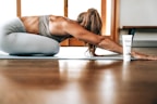 A person in athletic wear is performing a yoga pose on a mat indoors, with long hair falling forward. A tube of product labeled 'fringe mysa' is positioned nearby. The environment is well-lit with wooden flooring and a window in the background.