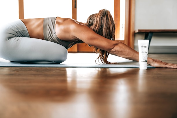 A person in athletic wear is performing a yoga pose on a mat indoors, with long hair falling forward. A tube of product labeled 'fringe mysa' is positioned nearby. The environment is well-lit with wooden flooring and a window in the background.