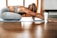 A person in athletic wear is performing a yoga pose on a mat indoors, with long hair falling forward. A tube of product labeled 'fringe mysa' is positioned nearby. The environment is well-lit with wooden flooring and a window in the background.