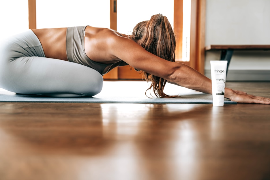 A person in athletic wear is performing a yoga pose on a mat indoors, with long hair falling forward. A tube of product labeled 'fringe mysa' is positioned nearby. The environment is well-lit with wooden flooring and a window in the background.