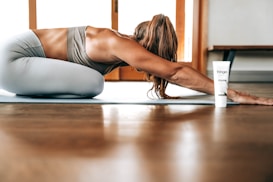 A person in athletic wear is performing a yoga pose on a mat indoors, with long hair falling forward. A tube of product labeled 'fringe mysa' is positioned nearby. The environment is well-lit with wooden flooring and a window in the background.