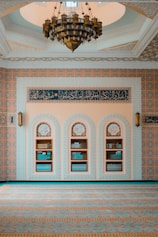 An intricately decorated room features a large chandelier hanging from a domed ceiling. The walls are adorned with ornate patterns and script in Arabic, showcasing elaborate geometric designs. There are three arched niches in the wall, each filled with books, indicating a scholarly or religious setting.