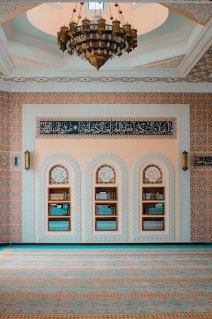 An intricately decorated room features a large chandelier hanging from a domed ceiling. The walls are adorned with ornate patterns and script in Arabic, showcasing elaborate geometric designs. There are three arched niches in the wall, each filled with books, indicating a scholarly or religious setting.