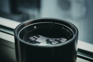 A large camera lens placed on a windowsill, reflecting some of the surrounding light. The lens has a matte black finish and visible branding or numbers around its rim. The background is softly blurred, suggesting a shallow depth of field.