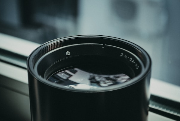 A large camera lens placed on a windowsill, reflecting some of the surrounding light. The lens has a matte black finish and visible branding or numbers around its rim. The background is softly blurred, suggesting a shallow depth of field.