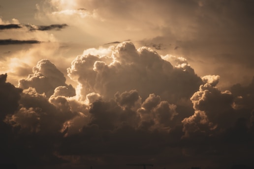 Large, dramatic clouds illuminated by sunlight, creating a contrast between the bright edges and the darker interiors of the clouds. The atmosphere appears moody and dynamic with variations in light and shadow across the cloud formations.