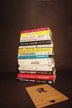 A stack of various books is neatly piled on a dark background. The titles of the books focus on topics such as business, personal development, leadership, and psychology. In front of the stack lies a wooden board with a letter 'R' engraved on it.