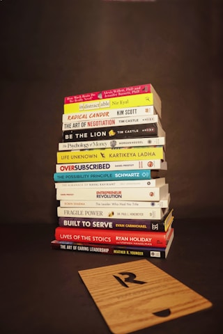 A stack of various books is neatly piled on a dark background. The titles of the books focus on topics such as business, personal development, leadership, and psychology. In front of the stack lies a wooden board with a letter 'R' engraved on it.