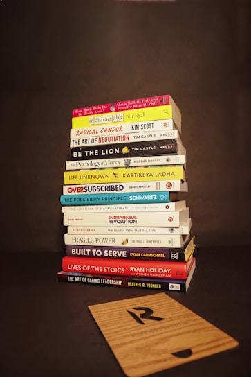 A stack of various books is neatly piled on a dark background. The titles of the books focus on topics such as business, personal development, leadership, and psychology. In front of the stack lies a wooden board with a letter 'R' engraved on it.