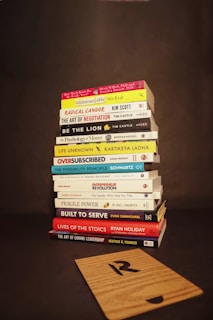 A stack of various books is neatly piled on a dark background. The titles of the books focus on topics such as business, personal development, leadership, and psychology. In front of the stack lies a wooden board with a letter 'R' engraved on it.