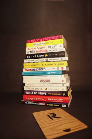 A stack of various books is neatly piled on a dark background. The titles of the books focus on topics such as business, personal development, leadership, and psychology. In front of the stack lies a wooden board with a letter 'R' engraved on it.