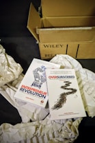 Two books by Daniel Priestley, titled 'Entrepreneur Revolution' and 'Oversubscribed', are placed on crumpled paper with an open cardboard box nearby labeled 'Wiley'.