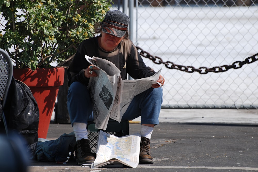 man in brown jacket and blue denim jeans sitting on red plastic chair reading newspaper