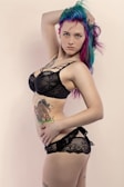 A person with vibrant, colorful hair featuring shades of blue, purple, and pink, posing in black lace lingerie. They have tattoos on their torso, including one of a cartoon horse. The background is a soft, neutral tone.