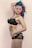 A person with vibrant, colorful hair featuring shades of blue, purple, and pink, posing in black lace lingerie. They have tattoos on their torso, including one of a cartoon horse. The background is a soft, neutral tone.