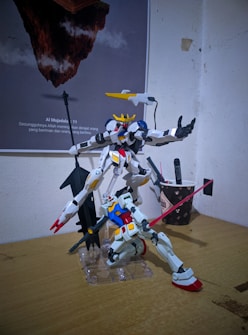Two intricately detailed Gundam model kits are positioned dynamically on a wooden surface. One model, featuring a predominantly white color scheme with blue and red accents, is kneeling with a pink beam saber held aloft. The other model stands upright, characterized by its bulkier design and a large weapon. In the background, there is a poster featuring floating rock islands with a quote that is partially visible. A small cup with writing utensils is also present on the desk.
