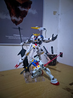 Two intricately detailed Gundam model kits are positioned dynamically on a wooden surface. One model, featuring a predominantly white color scheme with blue and red accents, is kneeling with a pink beam saber held aloft. The other model stands upright, characterized by its bulkier design and a large weapon. In the background, there is a poster featuring floating rock islands with a quote that is partially visible. A small cup with writing utensils is also present on the desk.