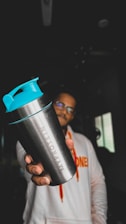 A person holding a metallic shaker bottle with turquoise lid prominently in the foreground. The figure is wearing a white hoodie with vibrant orange lettering. The background is dimly lit, with minimal visible details.