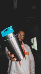 A person holding a metallic shaker bottle with turquoise lid prominently in the foreground. The figure is wearing a white hoodie with vibrant orange lettering. The background is dimly lit, with minimal visible details.