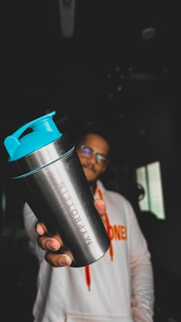 A person holding a metallic shaker bottle with turquoise lid prominently in the foreground. The figure is wearing a white hoodie with vibrant orange lettering. The background is dimly lit, with minimal visible details.