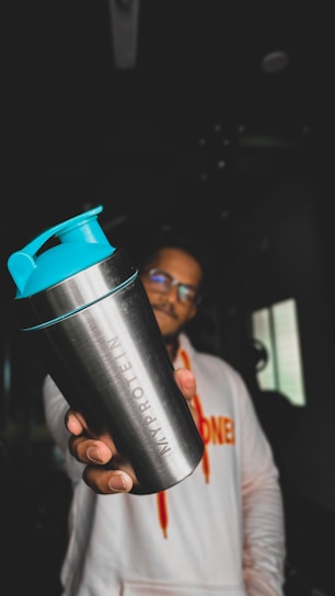 A person holding a metallic shaker bottle with turquoise lid prominently in the foreground. The figure is wearing a white hoodie with vibrant orange lettering. The background is dimly lit, with minimal visible details.