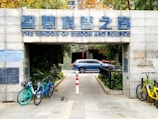 An entrance with a sign in both Chinese and English reading 'The Window of Wisdom and Science' above. Bicycles are parked on both sides of the entrance path, and a blue car is visible in the background beyond the entrance. The surroundings include green foliage and a paved pathway.