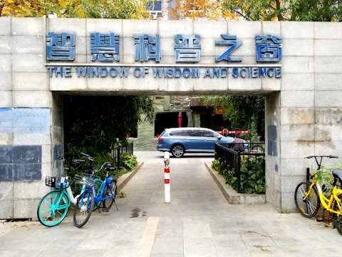 An entrance with a sign in both Chinese and English reading 'The Window of Wisdom and Science' above. Bicycles are parked on both sides of the entrance path, and a blue car is visible in the background beyond the entrance. The surroundings include green foliage and a paved pathway.