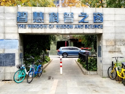 An entrance with a sign in both Chinese and English reading 'The Window of Wisdom and Science' above. Bicycles are parked on both sides of the entrance path, and a blue car is visible in the background beyond the entrance. The surroundings include green foliage and a paved pathway.