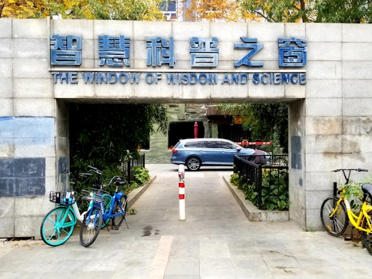 An entrance with a sign in both Chinese and English reading 'The Window of Wisdom and Science' above. Bicycles are parked on both sides of the entrance path, and a blue car is visible in the background beyond the entrance. The surroundings include green foliage and a paved pathway.
