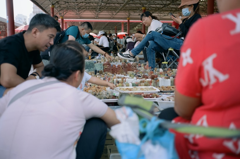 A busy market scene where several people are gathered around a stall displaying a variety of small items, possibly stones or carvings. The setting appears to be outdoors, under a wide structure with a roof. Some of the people are sitting or crouching, while others are standing and looking at their phones. There is a casual and lively atmosphere with interactions among the people.