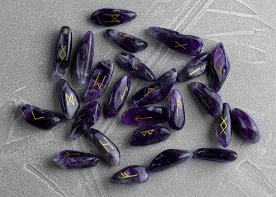 A collection of polished, purple amethyst stones with golden rune symbols engraved on them. The stones are scattered on a textured, gray surface, each stone displaying different rune markings.
