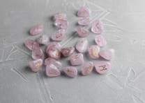 Several light pink crystals with engraved golden symbols are scattered on a textured gray surface. The stones have a polished finish, and the symbols seem to resemble ancient or mystical runes.