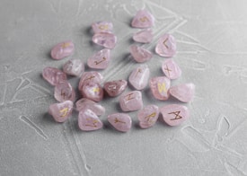 Several light pink crystals with engraved golden symbols are scattered on a textured gray surface. The stones have a polished finish, and the symbols seem to resemble ancient or mystical runes.