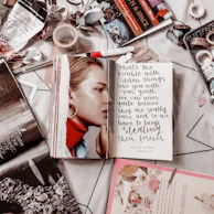 A cluttered desk with homemade junk journals, bright stickers, and a lit candle surrounded by pink bubblegum bubbles.