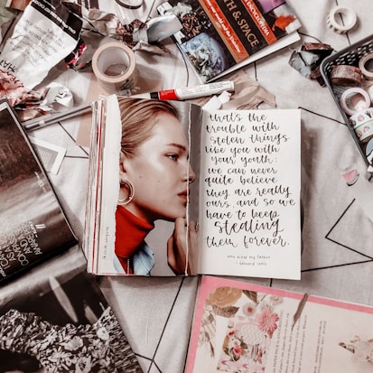 A cluttered tabletop featuring an open book with a black and white photograph of a woman on the left page, and a handwritten quote on the right page. Surrounding the book are various items, including tape, magazines, and art supplies like a red marker and washi tape. The surface appears to be covered with torn paper and bits of stationery.