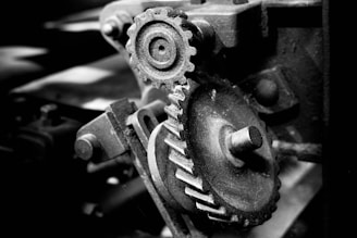 A close-up view of an intricate set of mechanical gears in monochrome. The gears have visible wear and are situated in a complex machine, with shadows highlighting their texture and details.