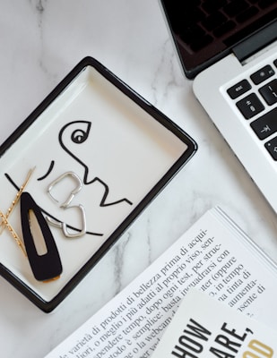 A close-up of a workspace scene featuring a laptop with a black keyboard partially visible. A decorative tray with abstract line art holds hairpins and metal clip pieces. Pages of a magazine with text in Italian are visible in the foreground. The surface is a sleek, white marble texture.