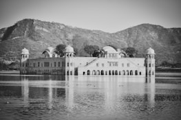 A large, ornate palace is situated in the middle of a body of water, surrounded by distant hills. The palace has multiple domes and arches, reflecting Mughal architectural style. The reflection of the palace is visible on the calm water surface, and there are a few birds visible in the water in front of the palace.