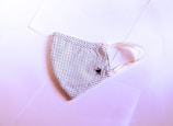 A fabric face mask with a black and white dotted pattern is laid flat on a slightly pink-tinted surface. The mask has elastic ear loops and a small embroidered crown detail on one side.