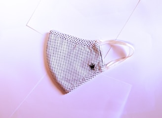A fabric face mask with a black and white dotted pattern is laid flat on a slightly pink-tinted surface. The mask has elastic ear loops and a small embroidered crown detail on one side.